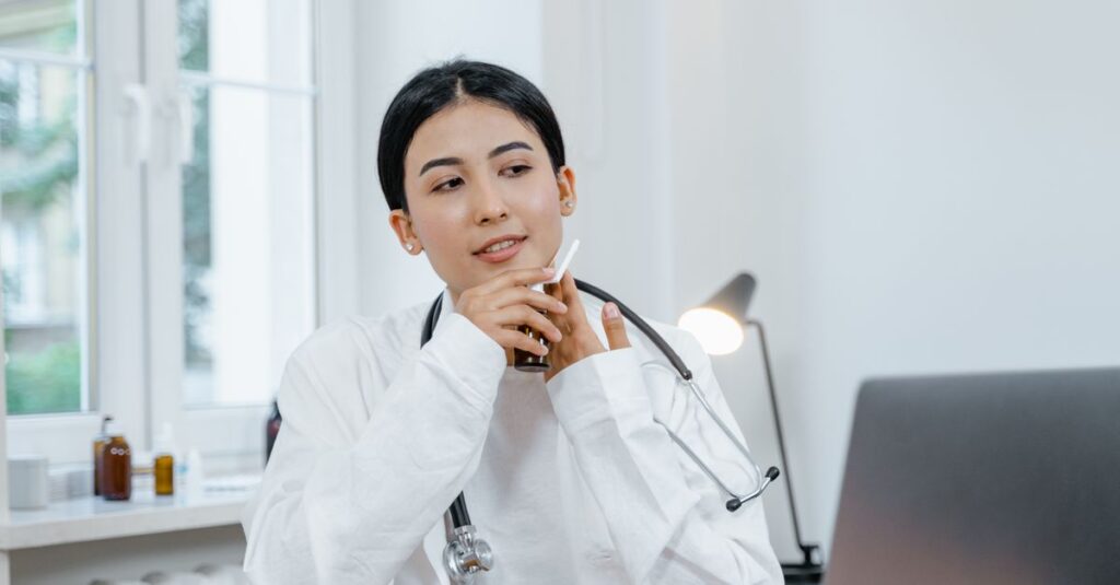 Asian female doctor using a smartphone for a video consultation, seated in a bright office setting. — in house vs hiring tele