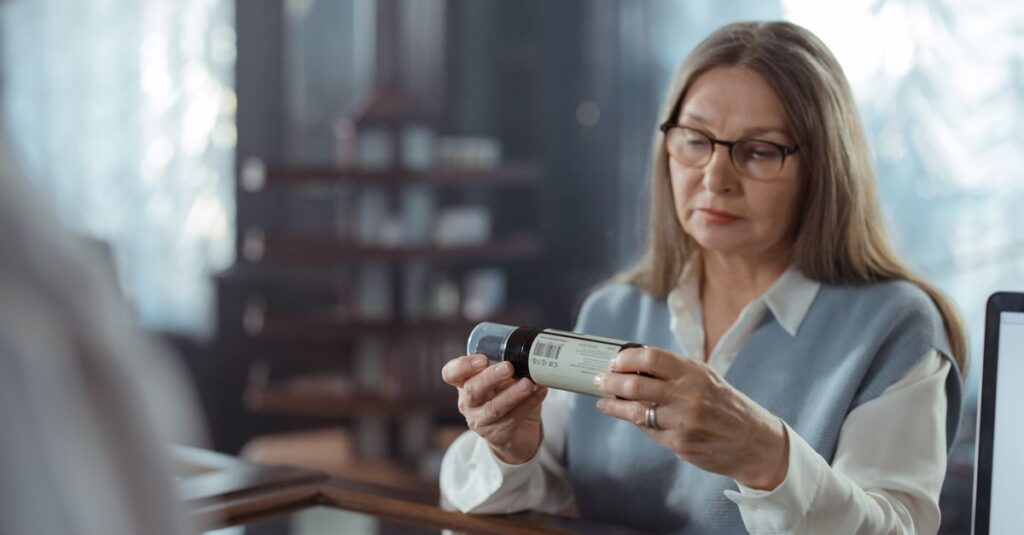 A senior woman with eyeglasses reviews a medicine bottle at a pharmacy counter indoors. — metformin weight loss